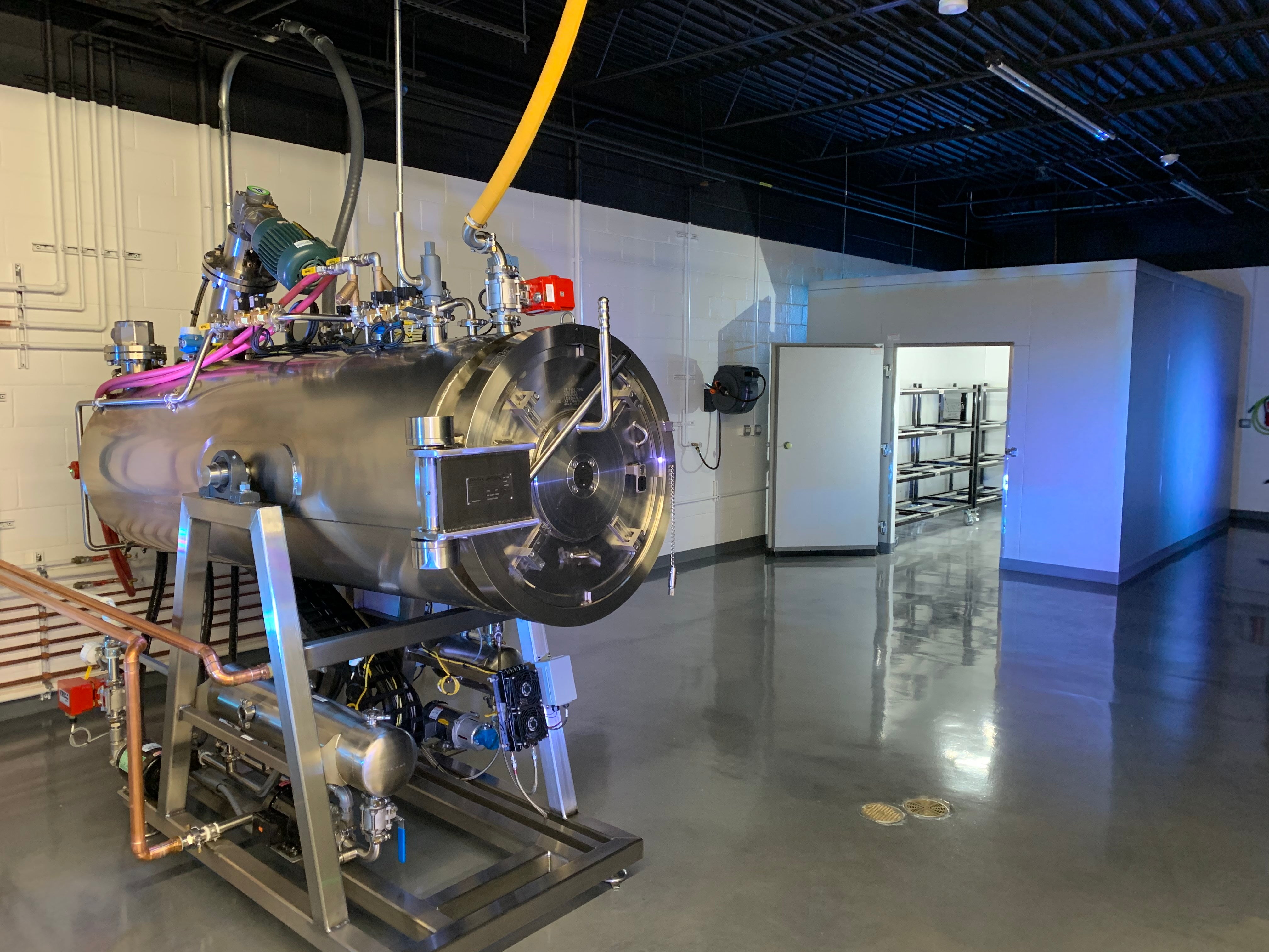 A stainless steel aquamation chamber positioned beside an American Mortuary Cooler equipped with mortuary body racks inside a clean, well-lit preparation room.